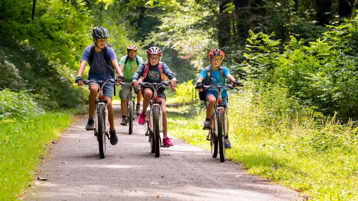 Three children on bikes on a traffic-free path of the National Cycle Network. Their grown up cycles behind. The path is surrounded by trees in full leaf. It is a warm, bright, sunny day.