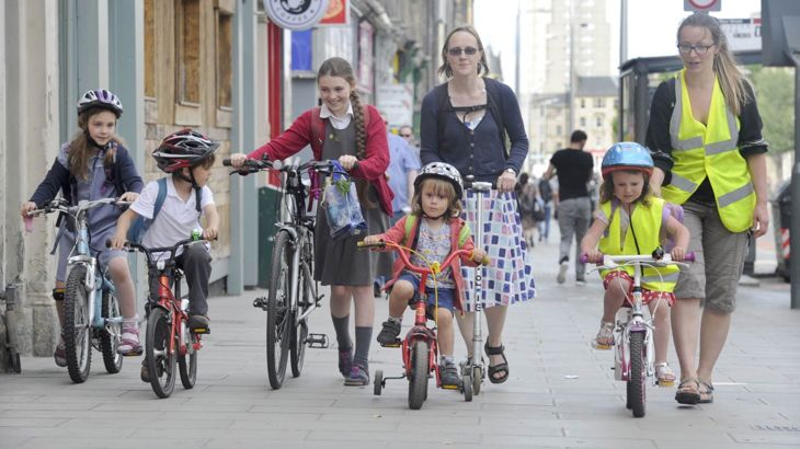 Young children cycling with adults walking on wide pavement