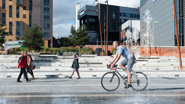 city plaza with fountains and people walking and cycling 