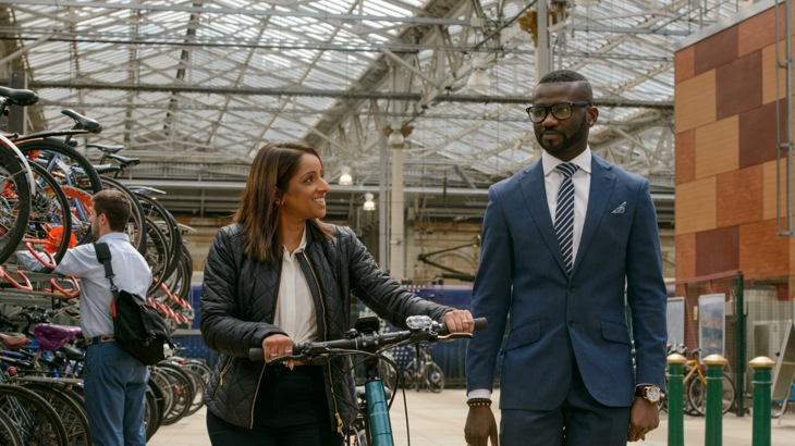 Two young professionals walking through a train station talking, one with a bicycle
