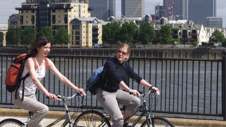 Two Women Cycling Along The Thames With Canary Wharf In The Background