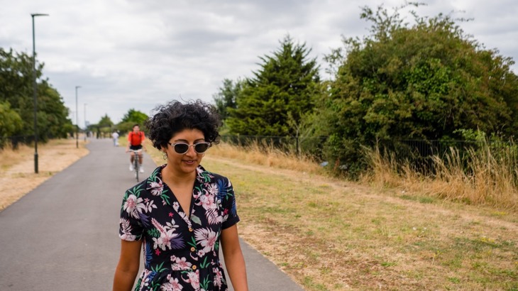 Woman in sunglasses walking on asphalt walking and cycling path with brown grass and green trees to the side and cyclist behind