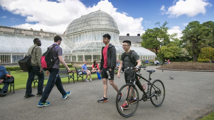 Two young men, one pushing a bike, are walking past The Palm House in Botanic Gardens, South Belfast on a sunny, blue sky and fluffy white cloud day. Other people are walking in the background and sat on benches enjoying the mild day.