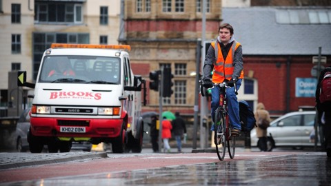 A man cycles in the rain in a city while wearing a high vis vest