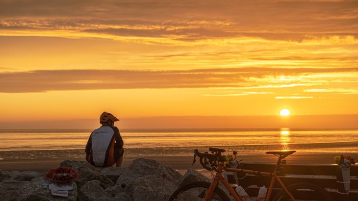 A cyclist sitting on some rocks looking out to sea as the sun sets, with poppy wreath and bike leaning up against a bench