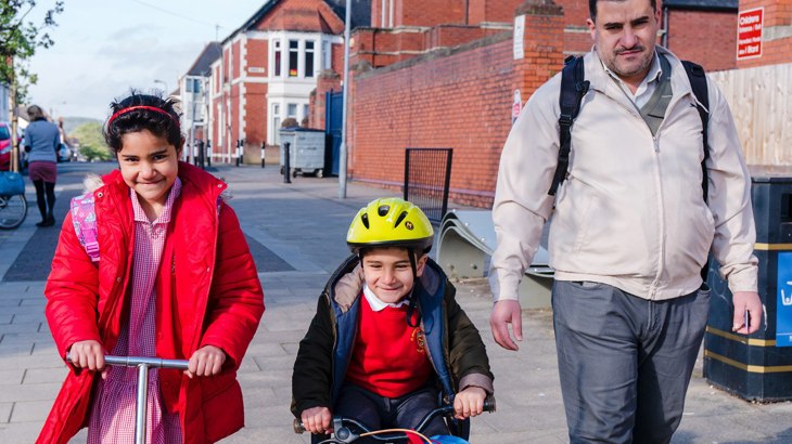 Family with dad walking, daughter scooting and son cycling to school together in the sunshine