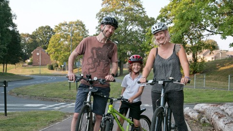Mum and dad with daughter in a park, wearing helmets and standing with their bikes smiling