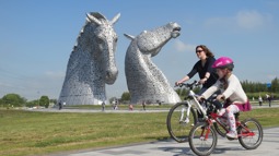 Young girl and woman riding cycles on path near giant horse head sculptures