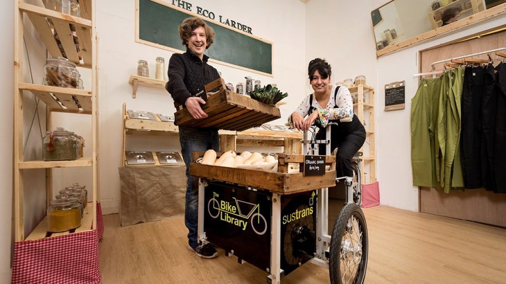 Man and woman standing inside a shop smiling next to a black, Walk Wheel Cycle Trust branded cargo bike they use for deliveries.