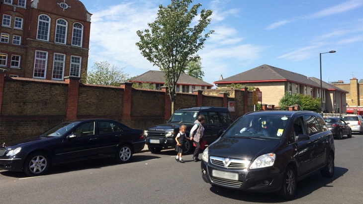 Adult and child holding hands trying to cross the street outside school with lots of cars driving down the road