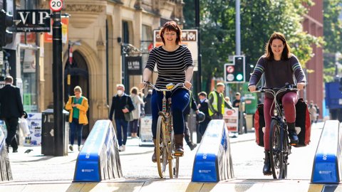 Two women smiling and chatting as they cycle together along a new temporary cycle lane through the high street.