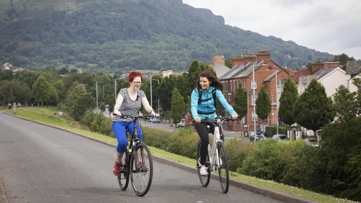 two women cycling along a traffic free path in the Water Works park, North Belfast with Cavehill mountain in the background