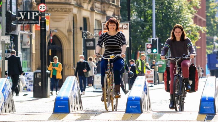 Two Women Cycling Through Barriers On Street In Manchester