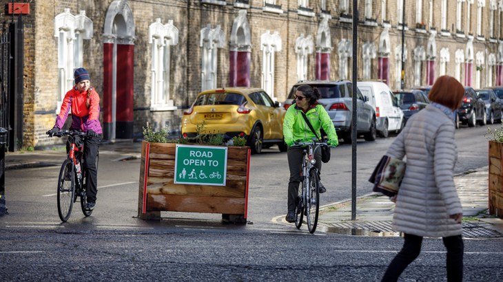 Two friends with bicycles stop at the end of a road next to a sign which reads "Road open to" followed by symbols for a parent and child, push scooter, wheelchair and bicycle.